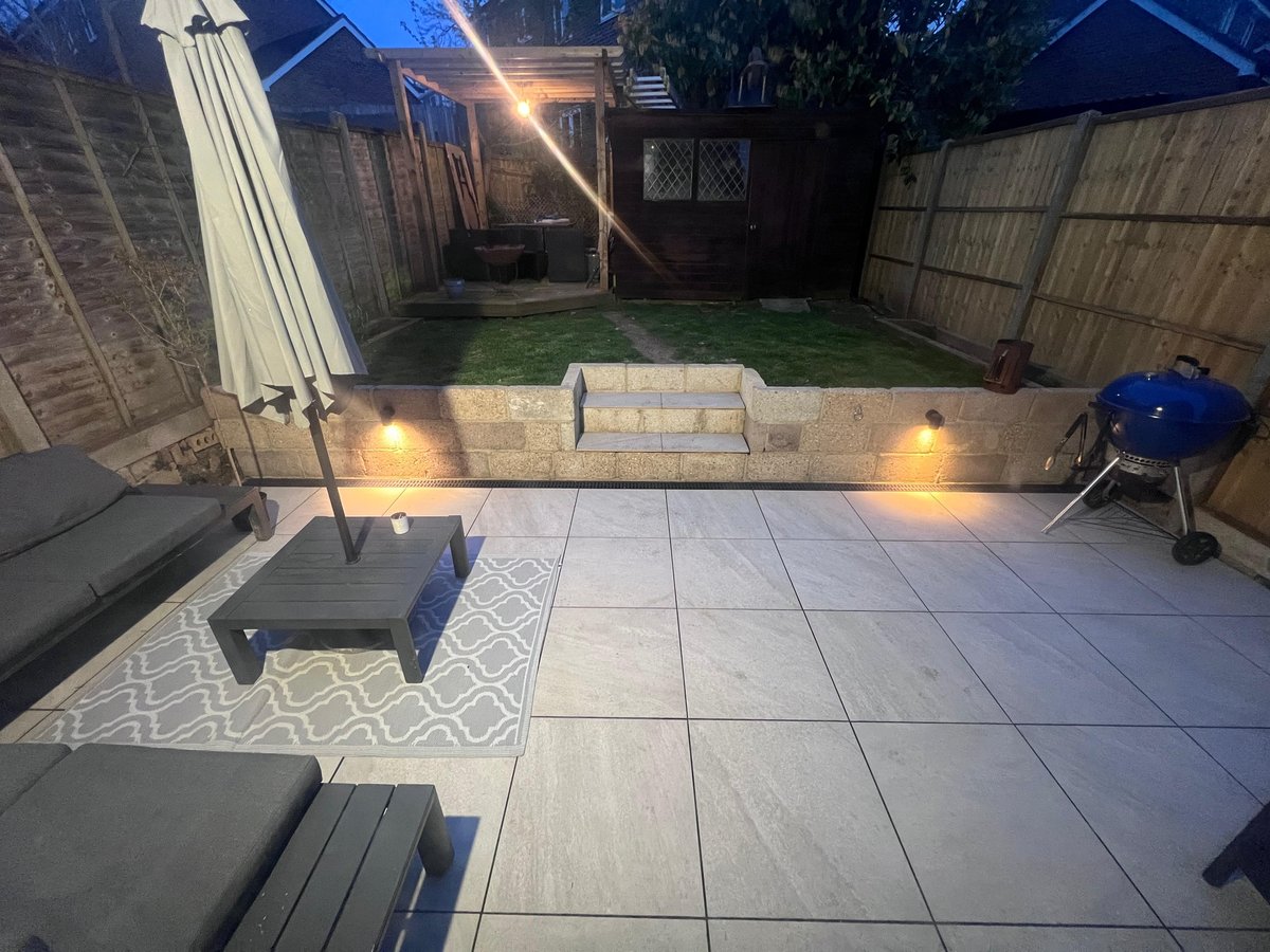 Landscaped back garden at dusk with grey porcelain patio, stone retaining wall with LED spotlights and rattan seating
